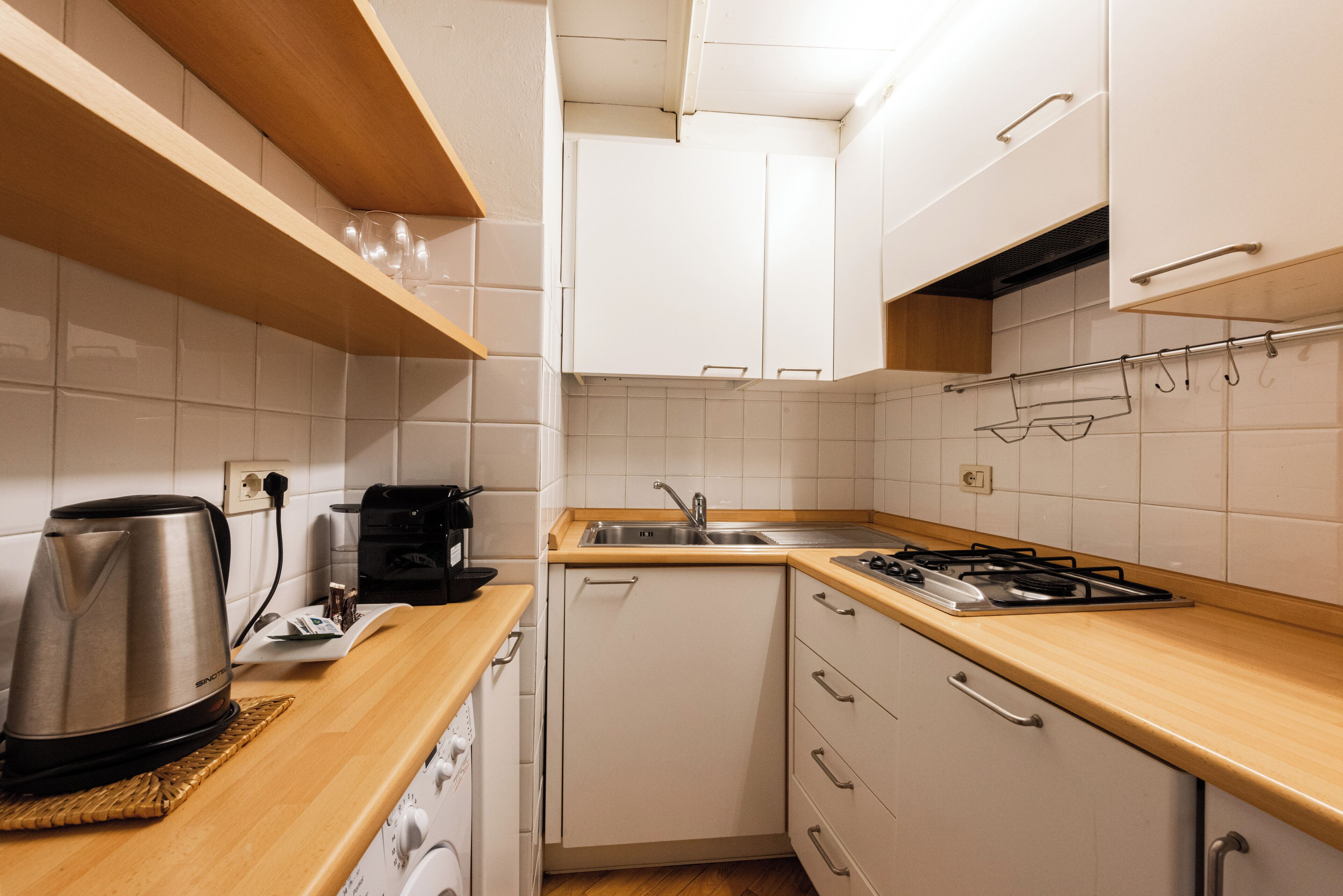 Et lille køkken med hvide skabe, en træbordplade, en stålkedel, kaffemaskine og en gasblus.