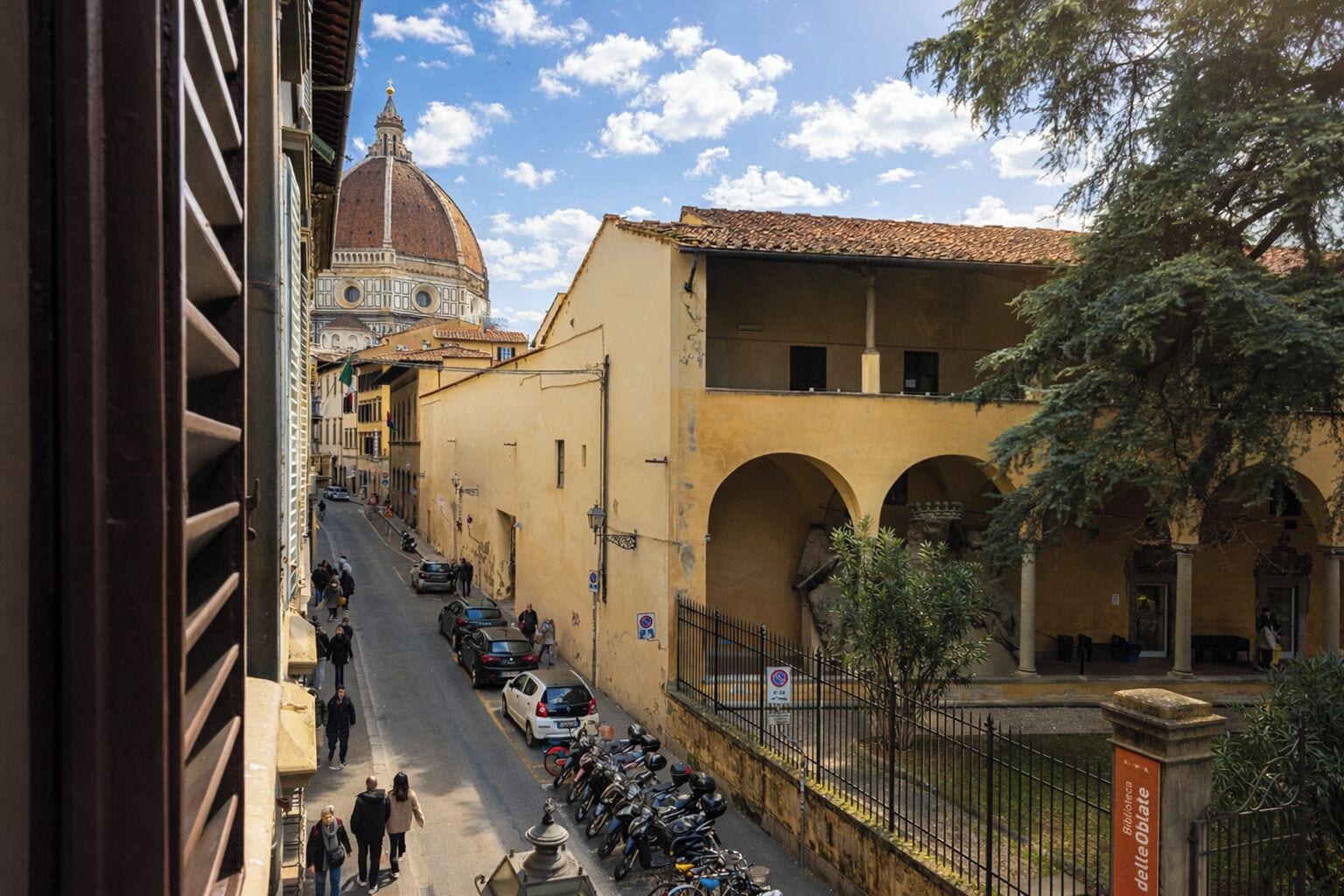 A street view with a prominent dome and yellow buildings, a narrow road with parked cars, and a pedestrian walkway.