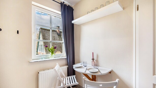 Une petite pièce avec une fenêtre, une table entourée de chaises, un radiateur et une étagère avec des bocaux.
