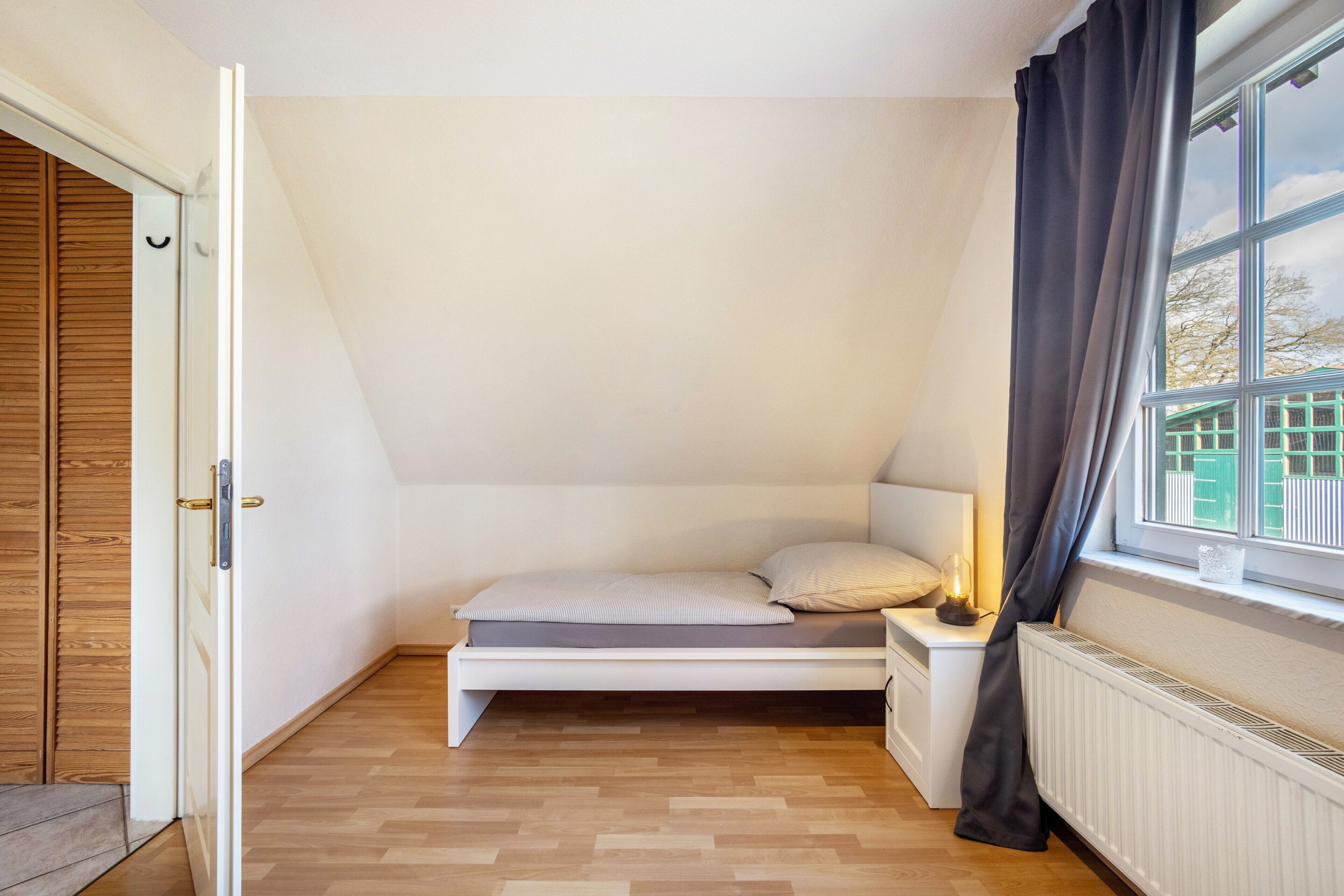 A bedroom with a bed, bedside table, window with curtains, wooden floor, and a door.