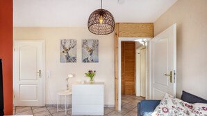 Une pièce comprenant une commode blanche, un bureau avec une lampe, un canapé agrémenté de coussins à motifs floraux et deux tableaux accrochés au mur.