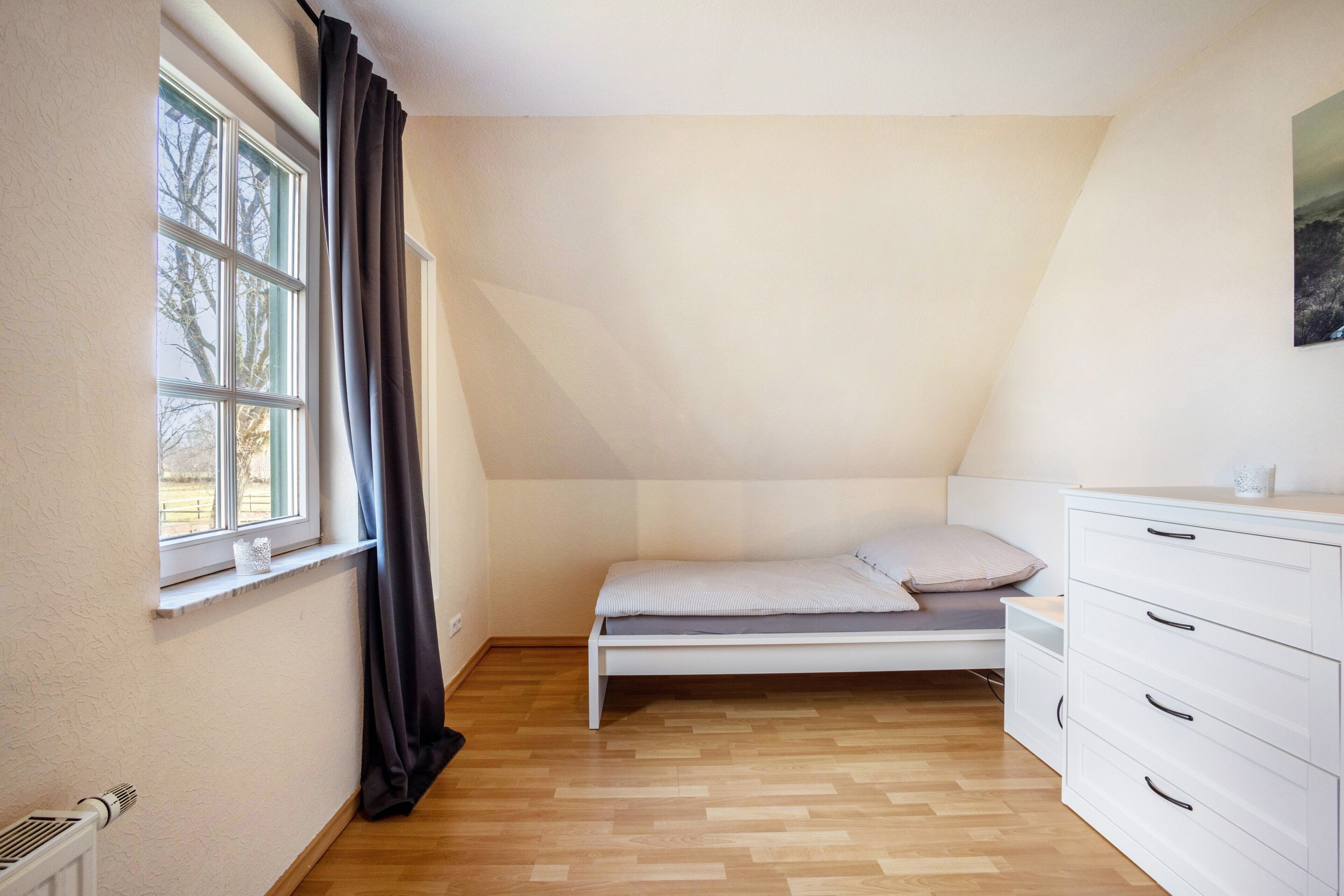 A small, single-bed room with a window, a radiator, and a white dresser.