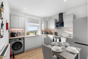 A modern kitchen with a built-in washing machine, a dining area with a table set for four, and a view of the outdoors through a window. - Birdies Nest Relax by the Links (Carnoustie)