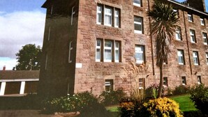 Un edificio de varios pisos con fachada de piedra, múltiples ventanas y un jardín bien cuidado con diversas plantas y flores.
