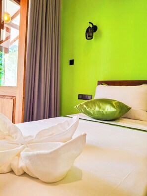 Un lit bien fait, avec du linge de lit blanc et un coussin vert, une tête de lit en bois, un luminaire fixé au mur et une fenêtre avec des rideaux.