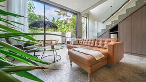 A modern living room with a leather sofa, a glass table, and a view of a tropical garden through large windows. - Pera Villa Ubud Bali. (Ubud)