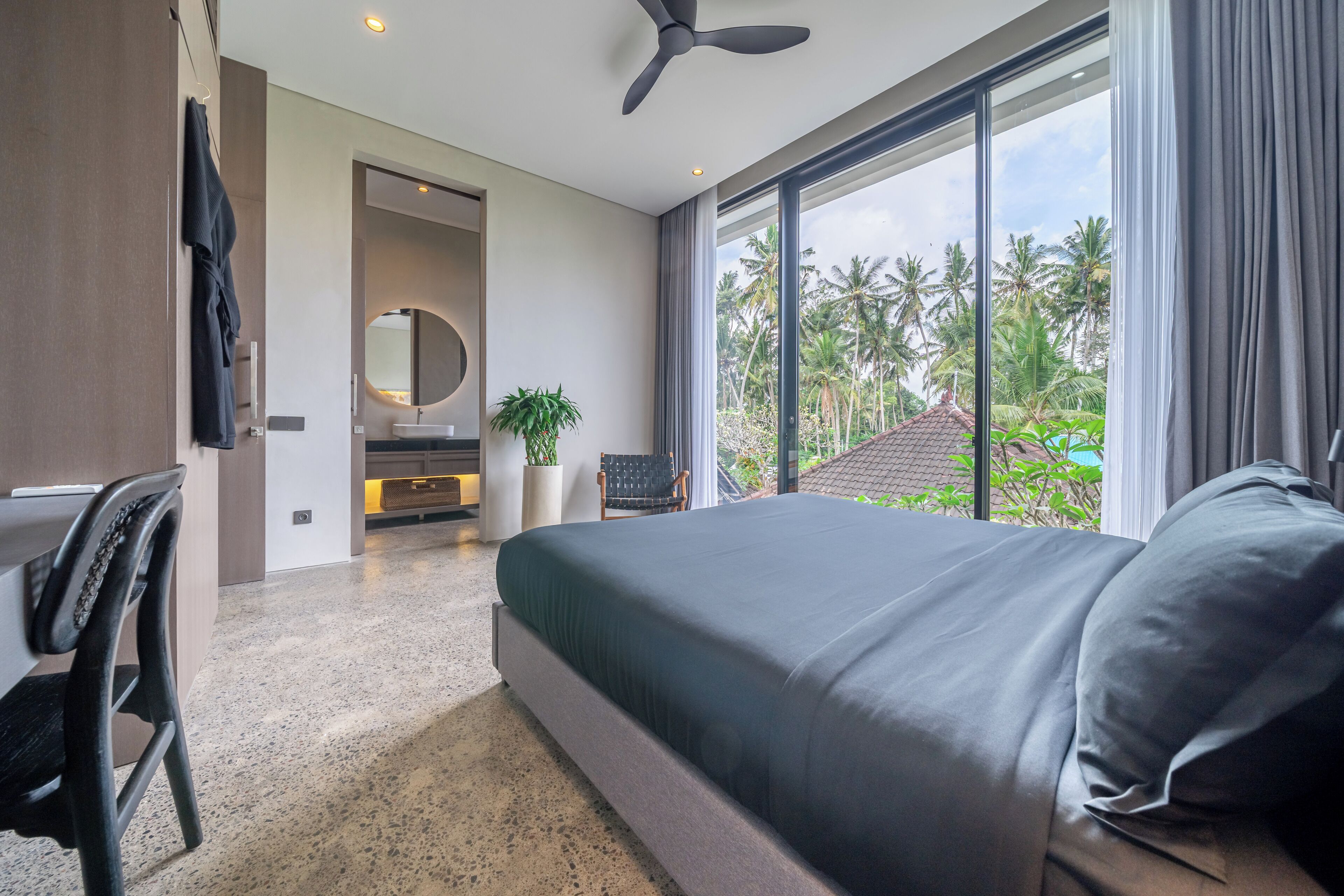 A modern hotel room with a large bed, a ceiling fan, a bathroom with a mirror, and a view of palm trees.