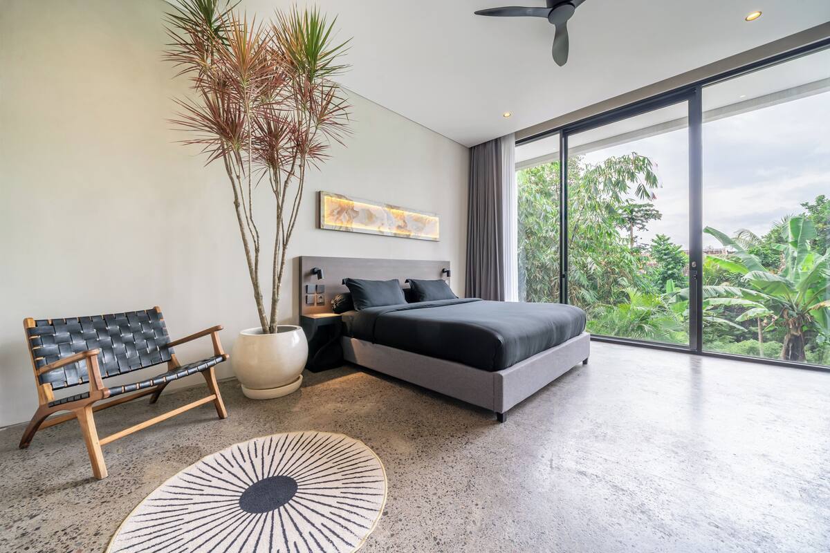 A modern bedroom with a large bed, a bench, a potted plant, and a view of greenery outside.