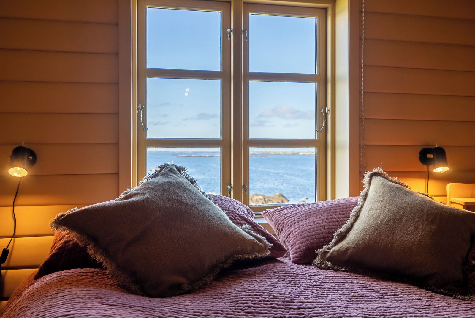 A bed with a view of the sea through a window, two bedside lamps, and a textured blanket.