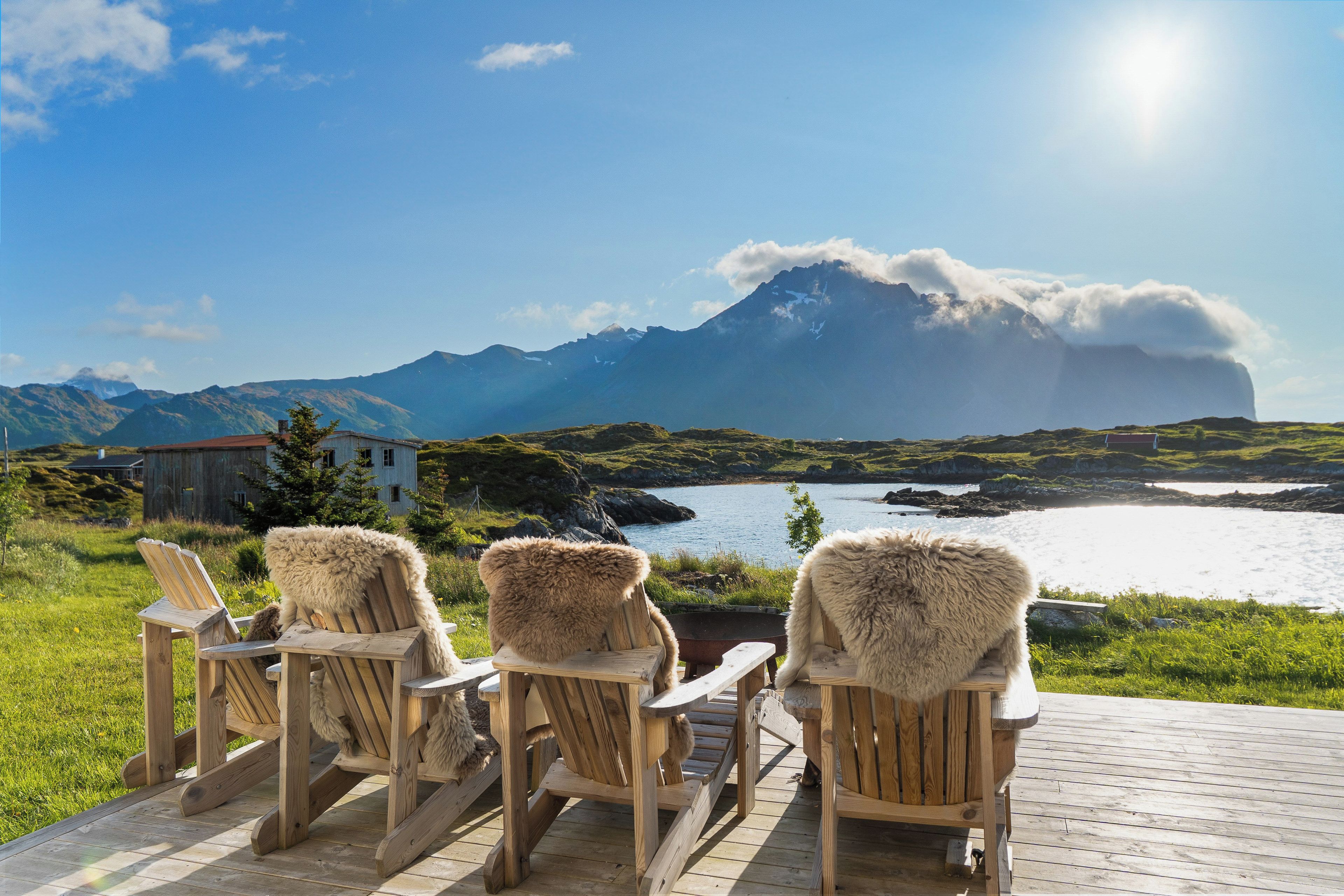 A wooden deck with four Adirondack chairs facing a body of water and mountains in the distance.
