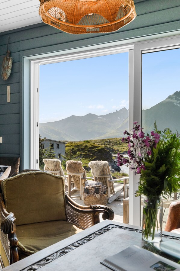 A room with a view of mountains, wicker furniture, and a table with a vase of flowers.