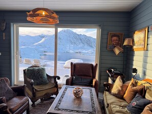 Une pièce chaleureuse avec une grande fenêtre donnant sur un paysage montagneux enneigé, un lustre en osier et un mélange de meubles en cuir et en tissu.