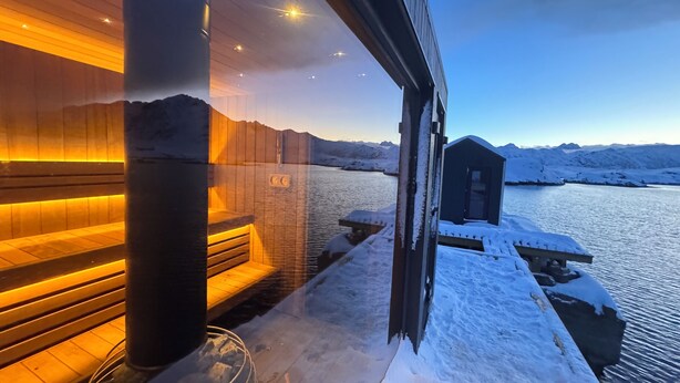 Zona de sauna con un banco de madera y una pileta de piedra, con vistas a un paisaje nevado y un edificio pequeño.