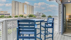 Un balcon avec des chaises bleues et une table, d’où l’on peut admirer un paysage urbain avec un grand immeuble et un ciel dégagé.