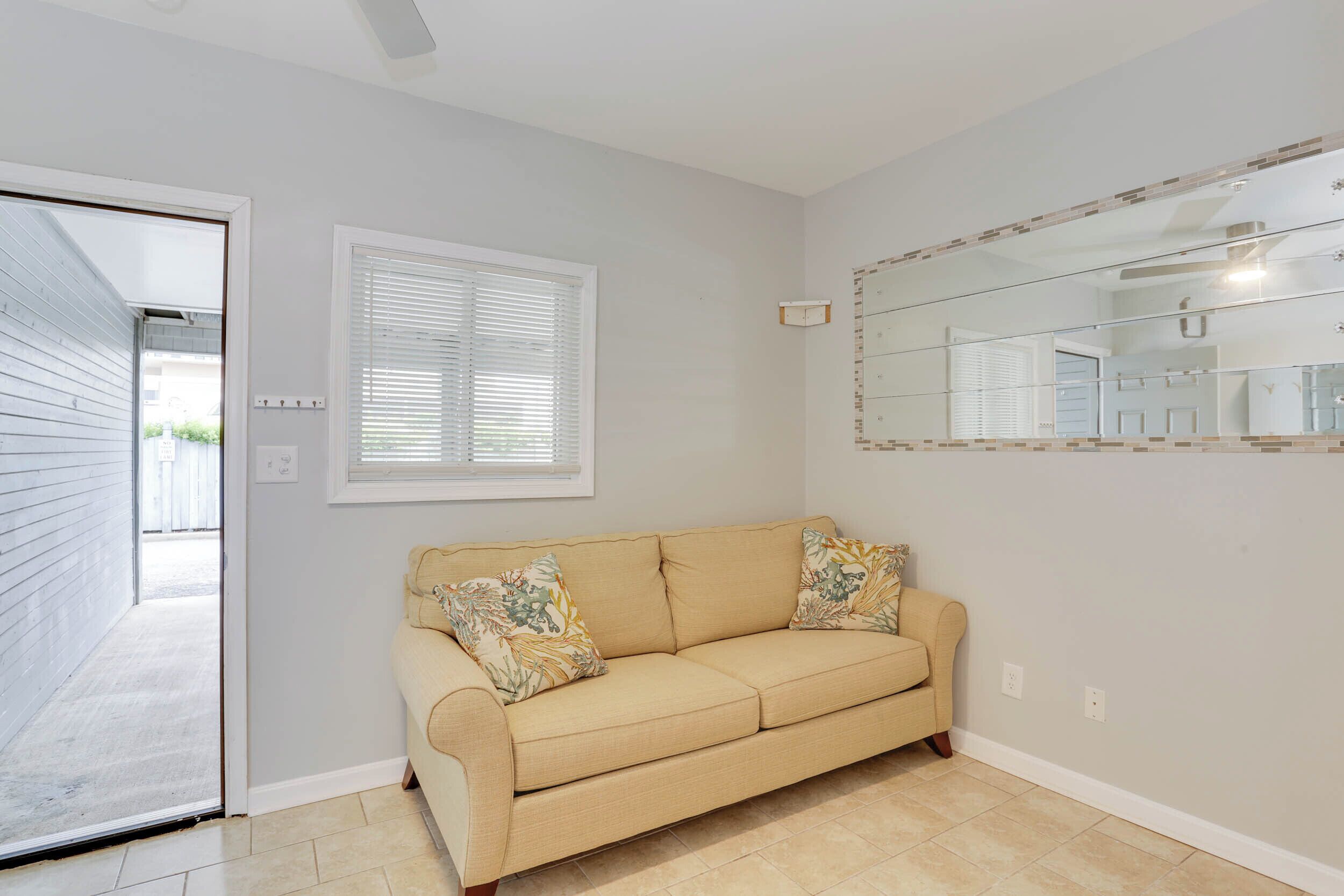 A room with a beige sofa, a window with blinds, a mirror, and a door leading outside.