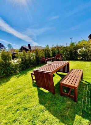 Une terrasse en bois avec une table et des bancs, entourée d’une pelouse bien entretenue et d’une végétation dense.