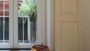 Une pièce avec une porte blanche, un panier en osier contenant des objets colorés, et une fenêtre donnant sur un balcon et des plantes.
