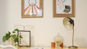 Un espace de travail comprenant un bureau blanc, une chaise blanche, une illustration d’ampoule encadrée, une affiche de l’alphabet encadrée, une lampe de bureau, une plante en pot et un certificat encadré.