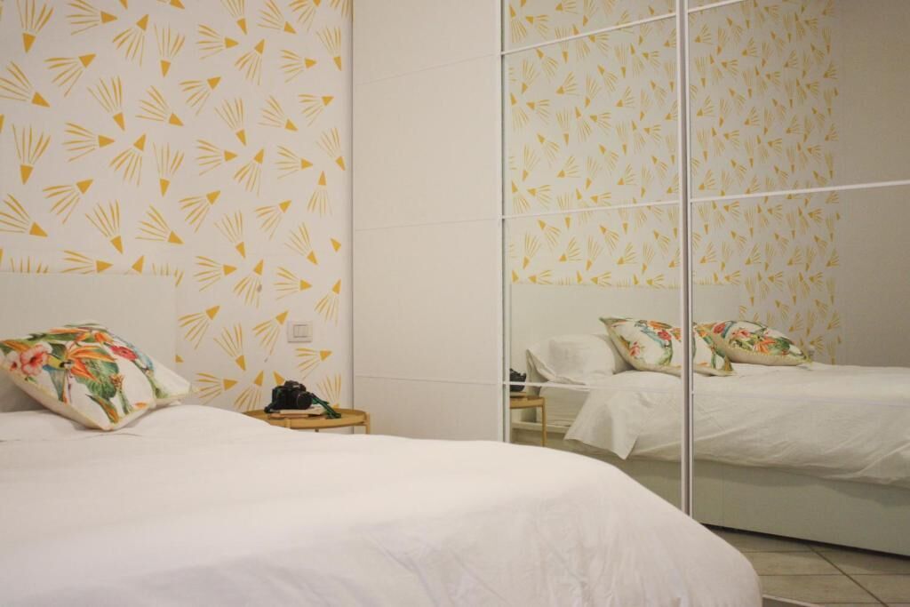 A bedroom with a mirrored wardrobe, a bed with floral pillows, and a small bedside table with a camera.