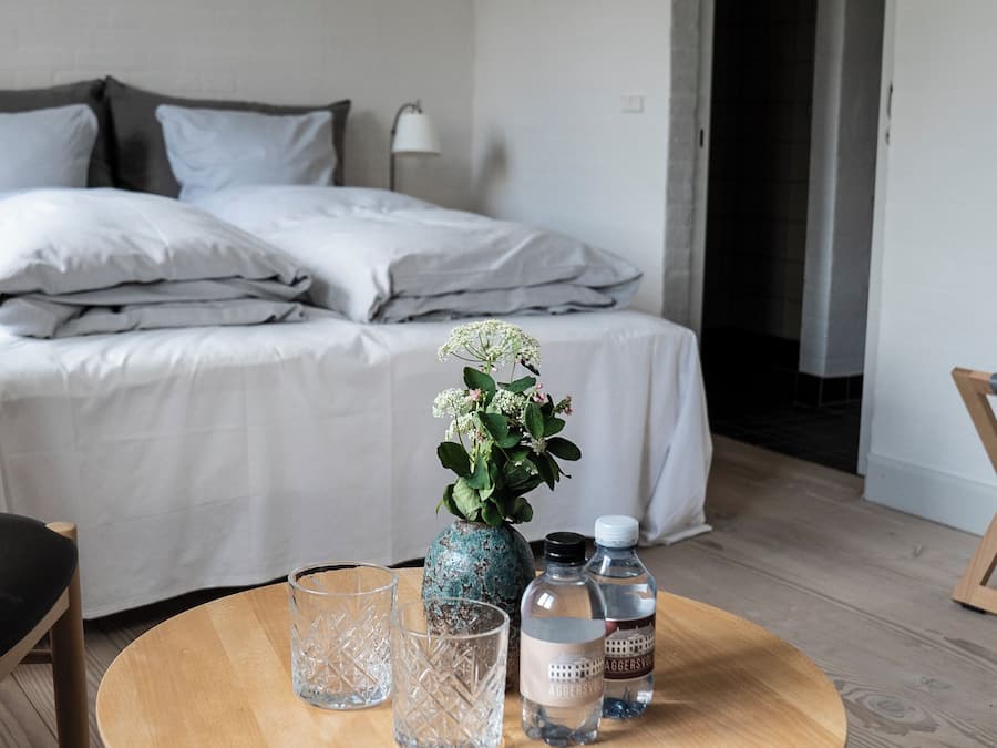 A neatly made bed with pillows, a bedside table with a vase and water bottles, and a chair with a cushion.