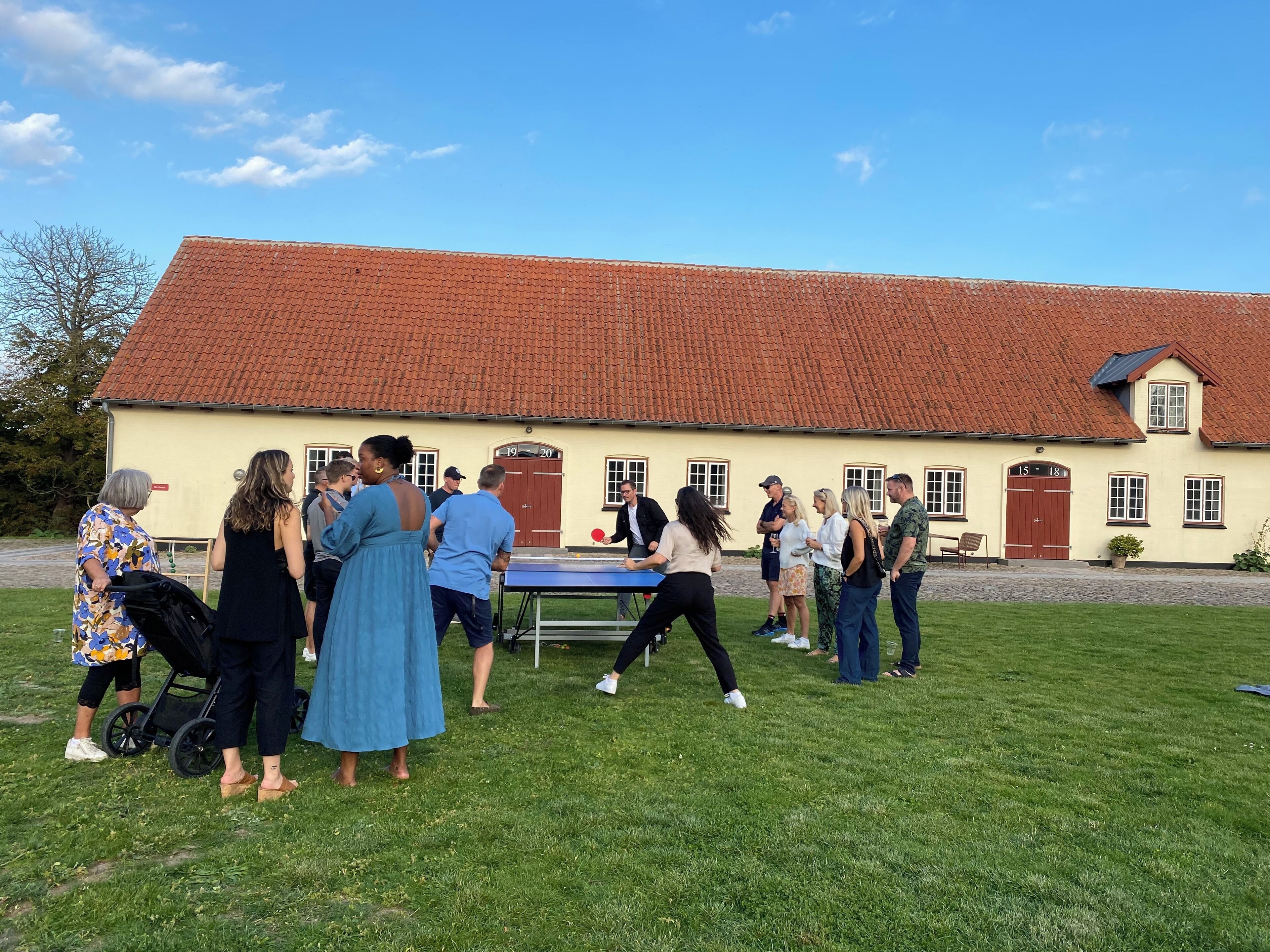 A group of people playing table tennis outdoors in front of a building with a red-tiled roof and numbered doors.