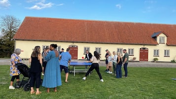 A group of people playing table tennis outdoors in front of a building with a red-tiled roof and numbered doors.