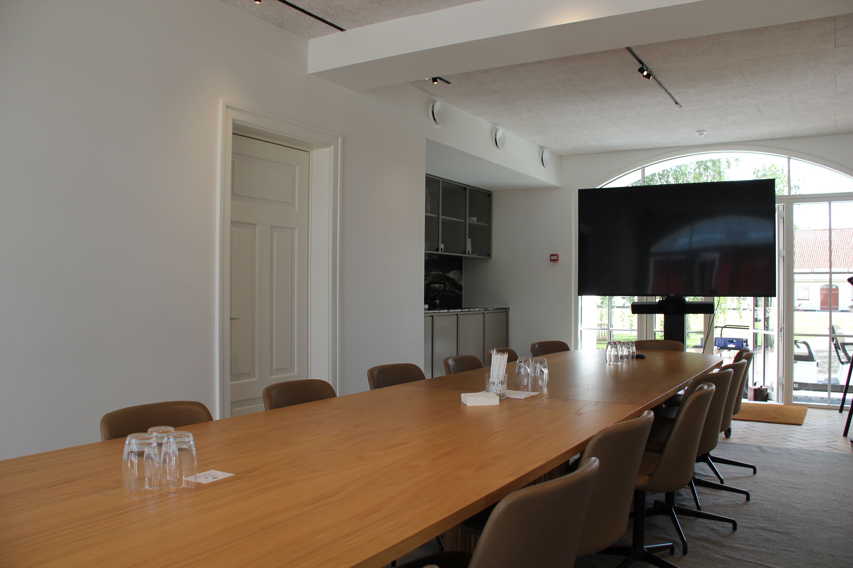 A conference room with a long table, chairs, a flat-screen TV, and a door.