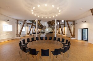 A spacious room with a circular arrangement of chairs, wooden beams, and a high ceiling with hanging lights.