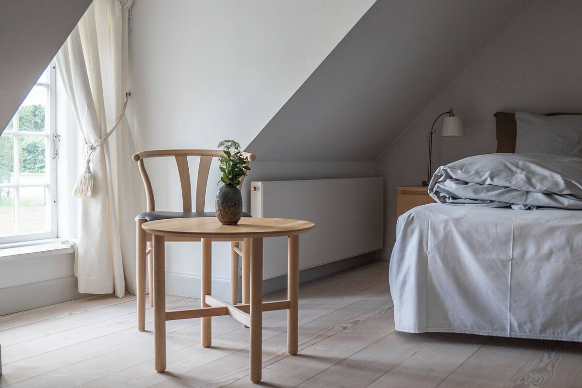 Una camera da letto con soffitto spiovente, un letto con lenzuola grigie, una sedia di legno con una pianta e un tavolino.