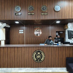 A reception desk with clocks indicating Japan, New York, and New Delhi times, a sign for advance payment, and a logo with a crown and laurel wreath. - Ruchi Resort (Hisar)