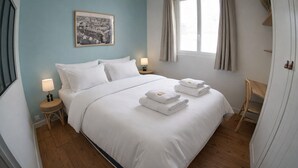 Een netjes opgemaakt bed met witte lakens en handdoeken, een klein nachtkastje met een lamp, een ingelijste foto aan de muur, en een raam met gordijnen.