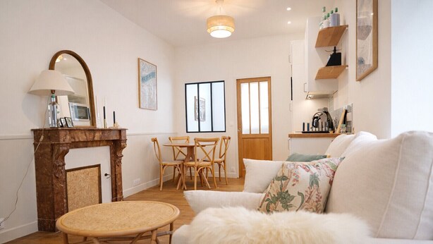 Een woonkamer met een witte bank, een rond rieten tafeltje, een houten salontafel, een open haard en een eethoek met houten stoelen en een houten tafel.