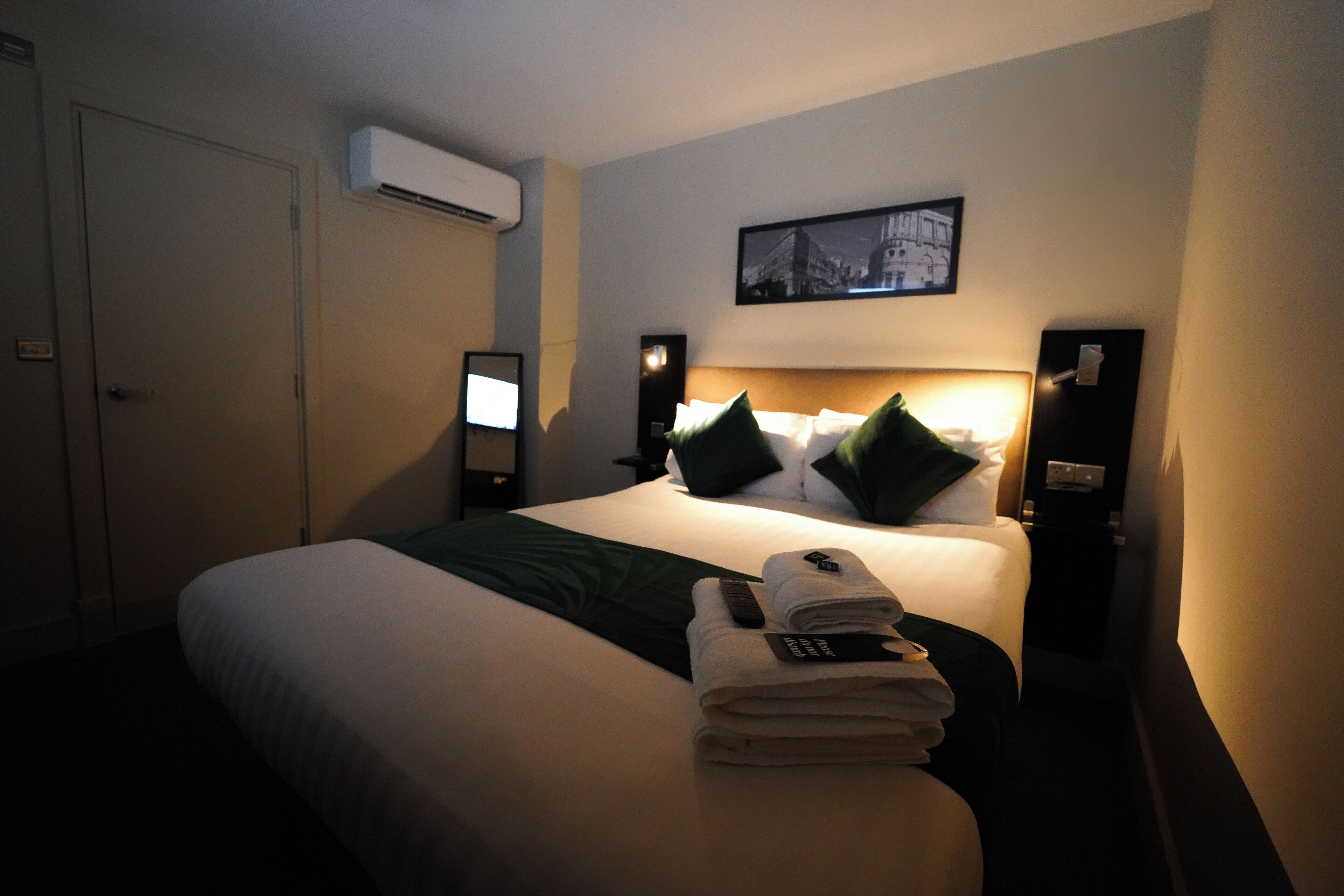 A hotel room with a neatly made bed, a bedside table with a lamp, a wall-mounted air conditioner, and a framed picture above the bed.