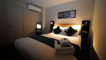 A hotel room with a neatly made bed, a bedside table with a lamp, a wall-mounted air conditioner, and a framed picture above the bed.