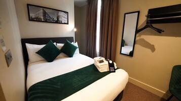 A hotel room with a neatly made bed, a bedside table with a phone and a bottle, a mirror, and a picture on the wall.