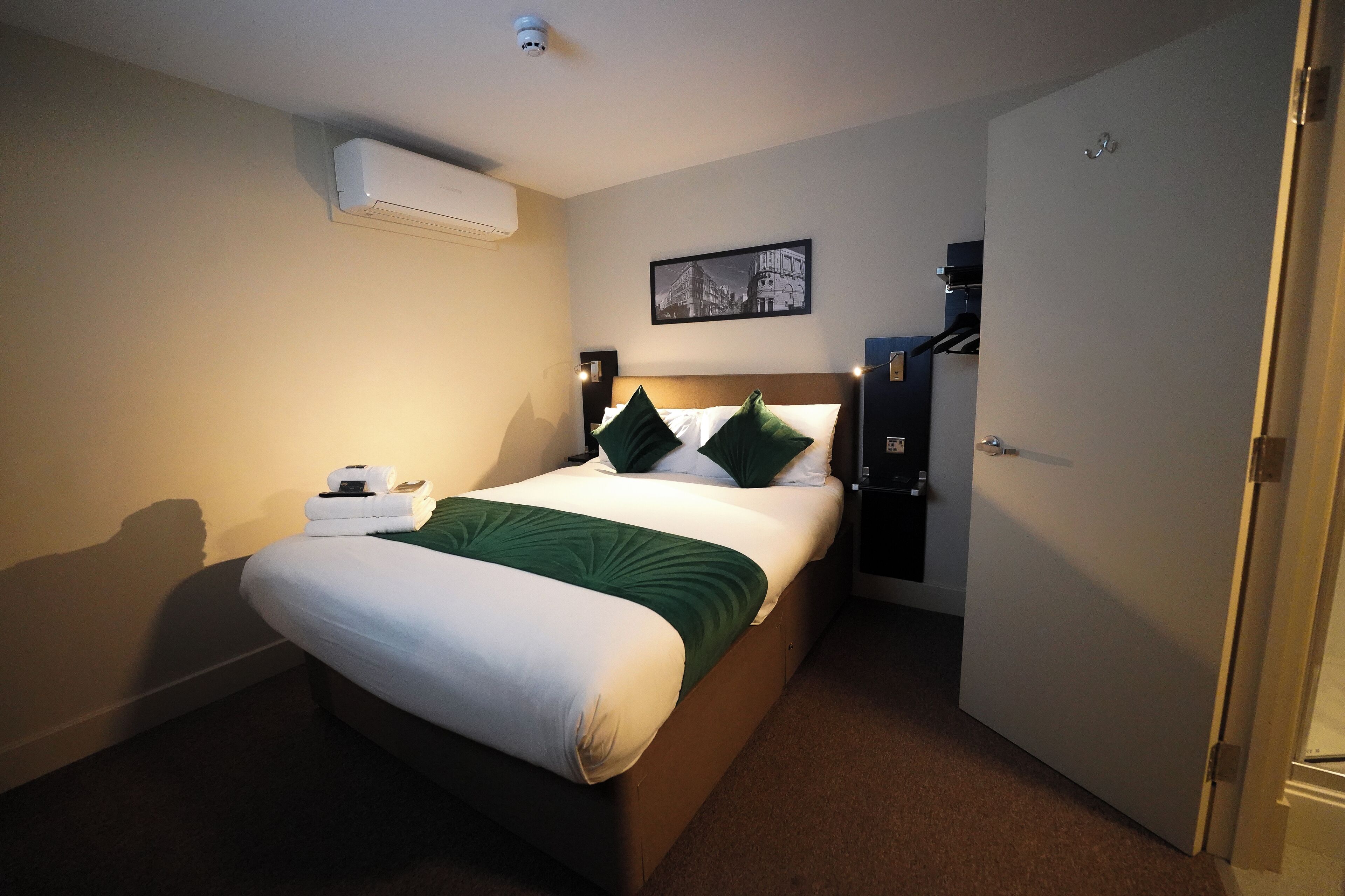 A hotel room with a bed, a nightstand, an air conditioning unit, a door, and a wall-mounted picture.