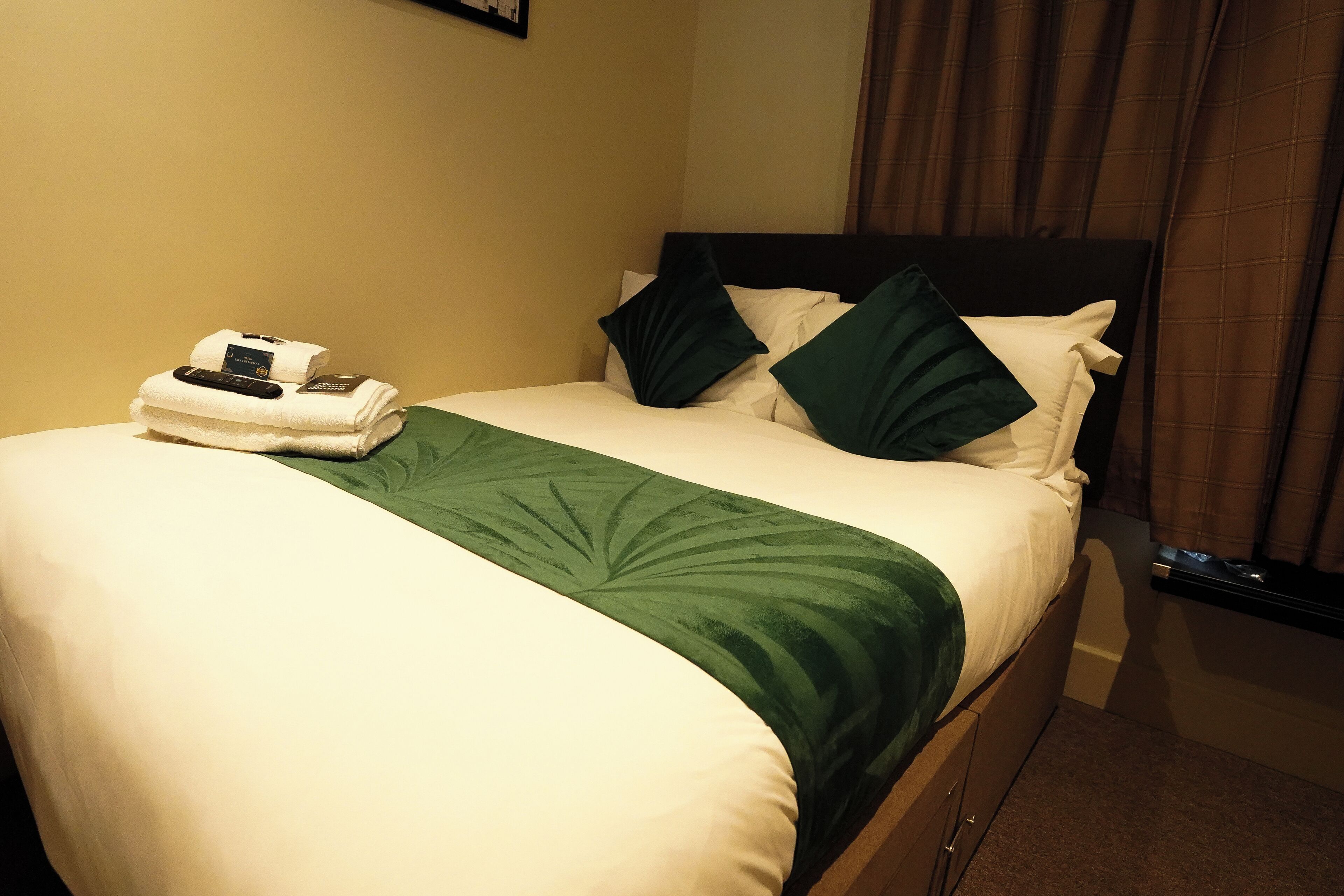 Ein ordentlich bezogenes Bett mit einer grünen Decke, weißen Kissen und einer Fernbedienung auf einem gefalteten Handtuch.