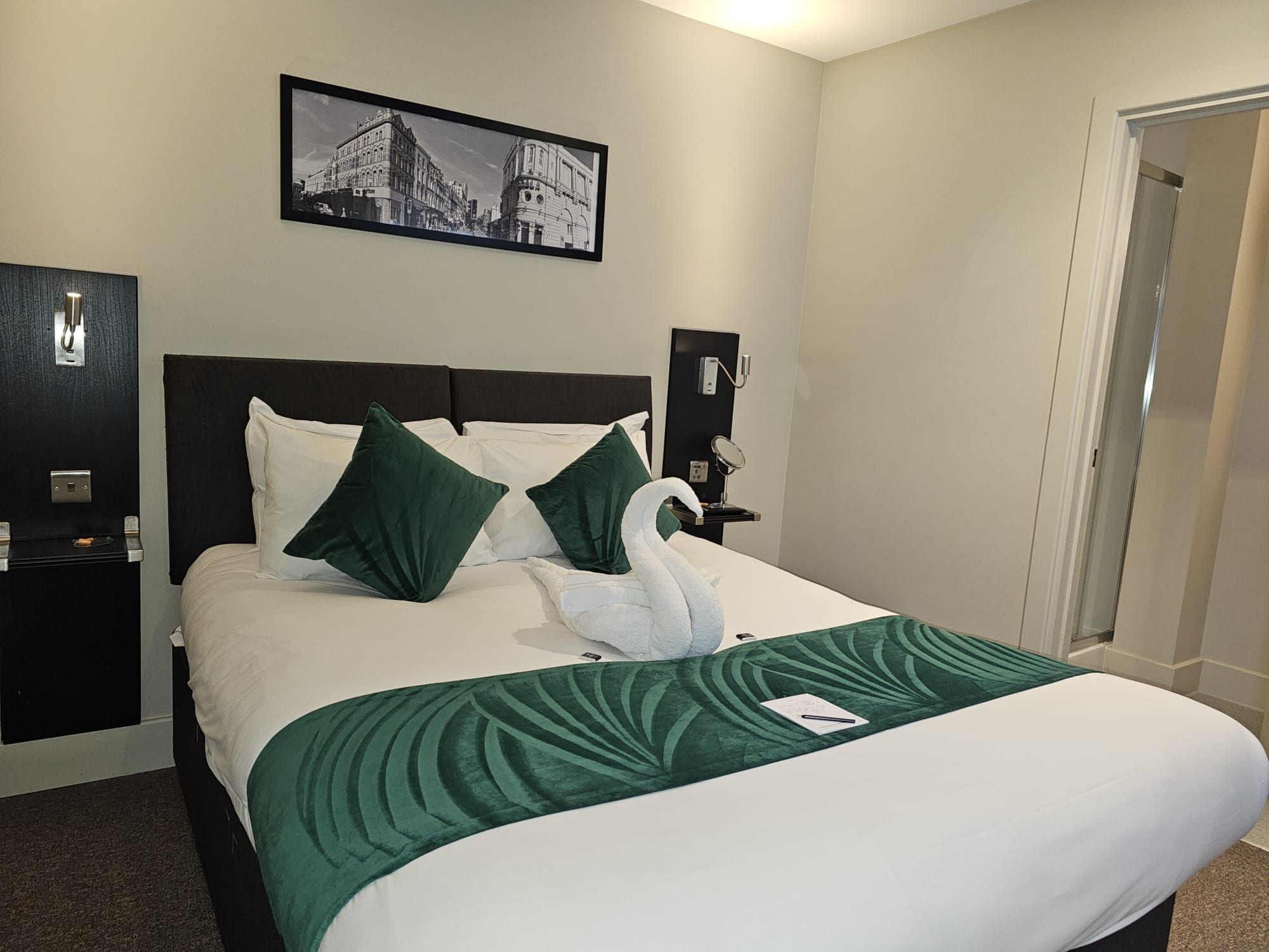 A neatly made bed with a swan-shaped pillow and a green patterned bedspread in a hotel room.