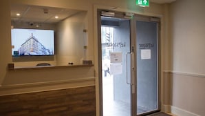 En hotellentrén med en glasdörr, en receptionsdisk och en digital skärm som visar en bild av byggnaden.