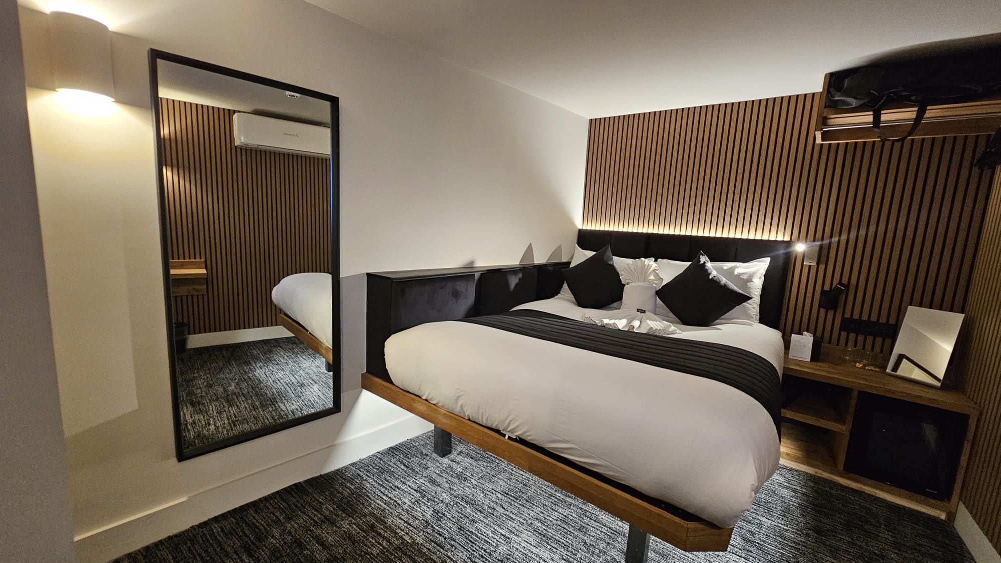 Ein modernes Hotelzimmer mit einem großen Bett, einem Nachttisch, einem Spiegel und einer Wand-Klimaanlage.