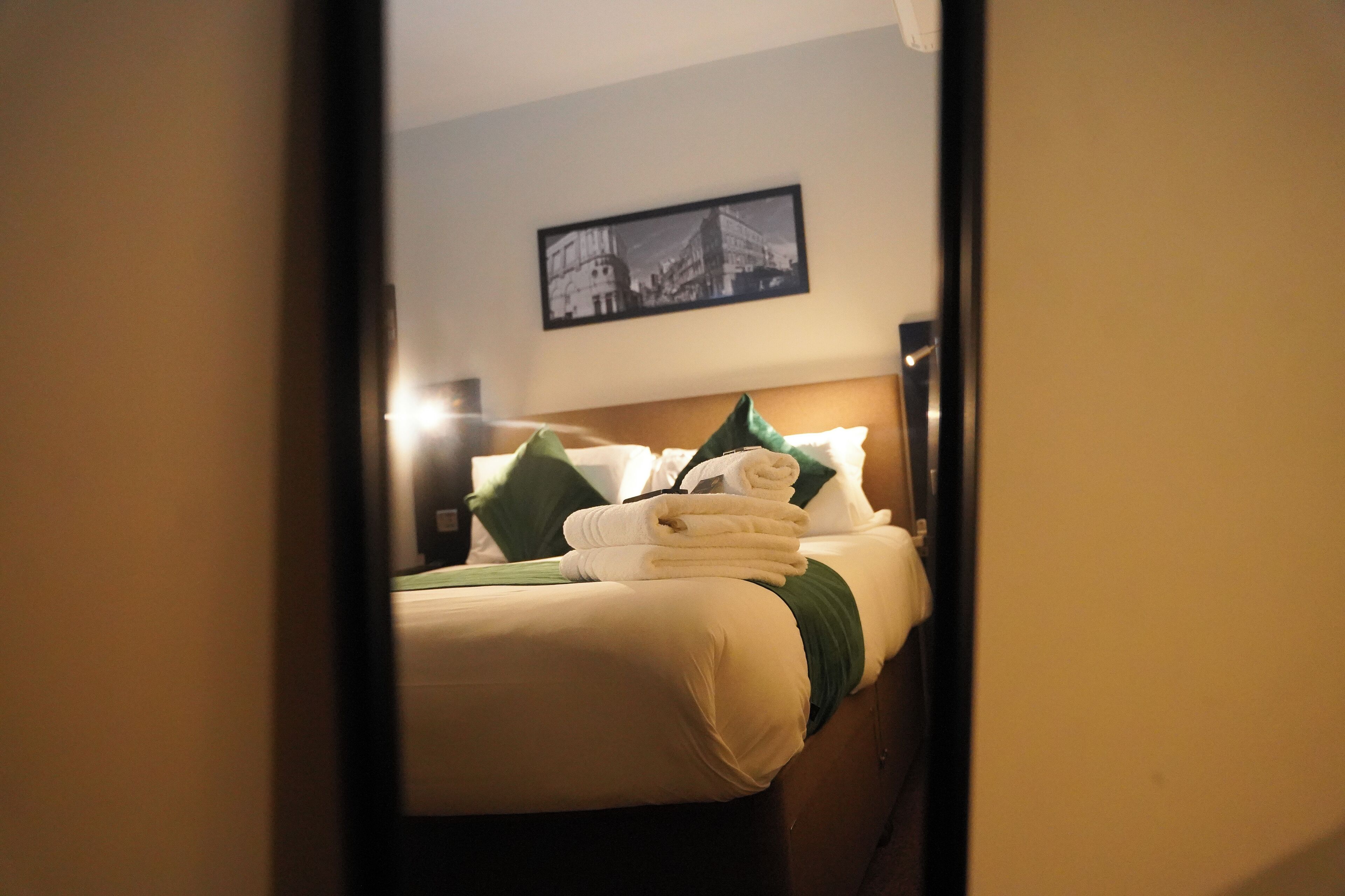 A hotel room with a neatly made bed, a framed picture on the wall, and a visible door frame.