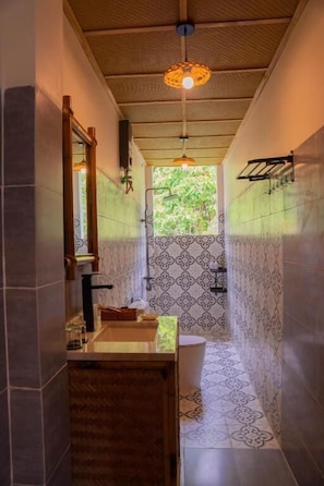 Um banheiro estreito com azulejos estampados, toucador de madeira, luminária pendente e janela com vista para o verde.