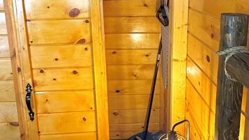 Interior de una cabaña de madera con una puerta corredera, una escoba, un farolillo y una pequeña mesa con una planta.