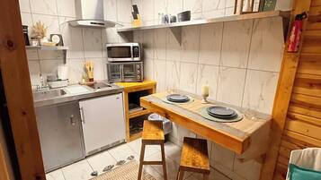 Cocina compacta con microondas, tostadora y lavabo. Hay un taburete de madera y una mesita pequeña con platos.