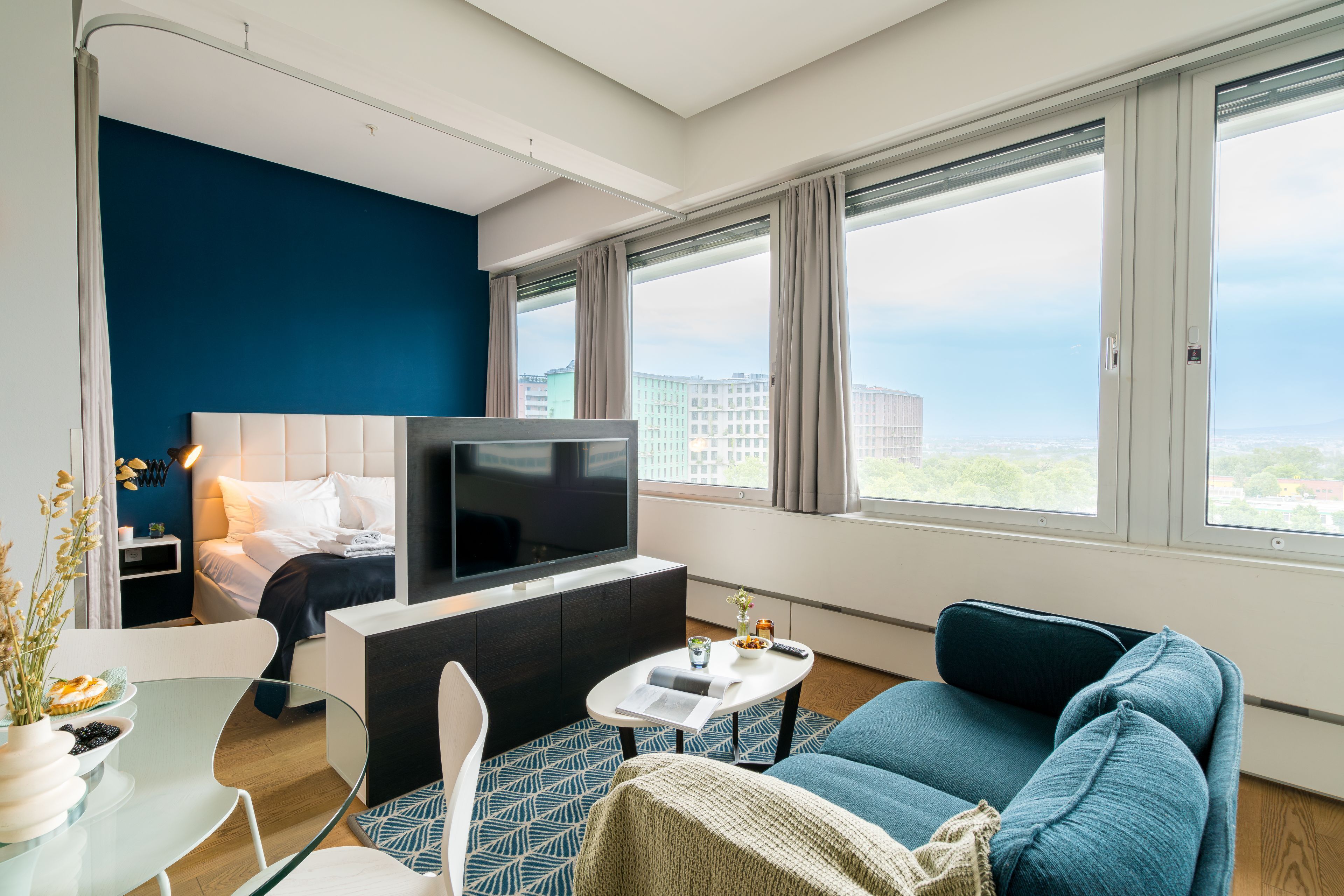 A modern hotel room with a bed, a TV, a sofa, a small table, and large windows offering a city view.
