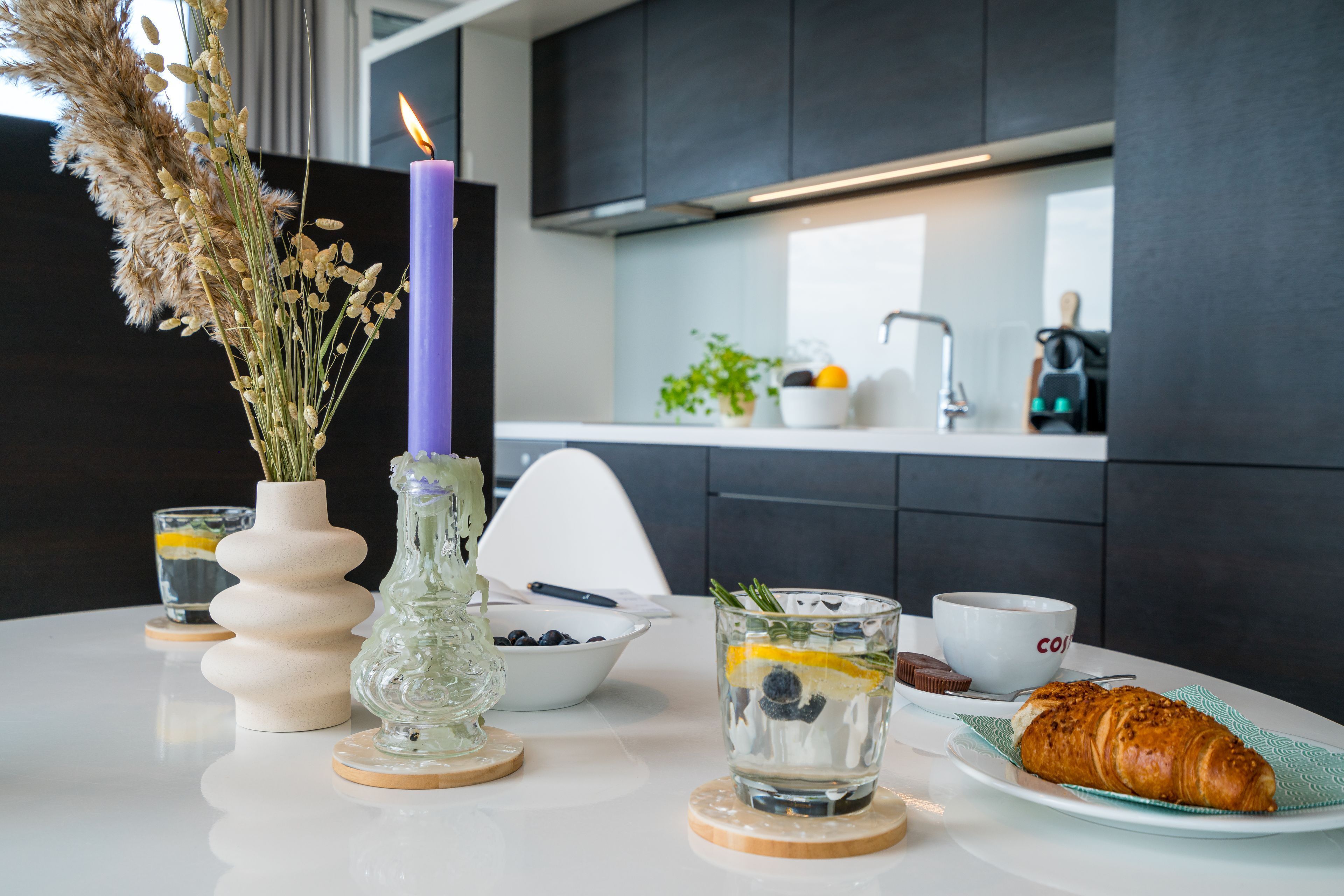 Una cocina moderna con armarios oscuros, una encimera blanca, un florero con plantas secas, una vela encendida, un cuenco de arándanos, un vaso de agua con limón y menta, una taza de café y un croissant en un plato.