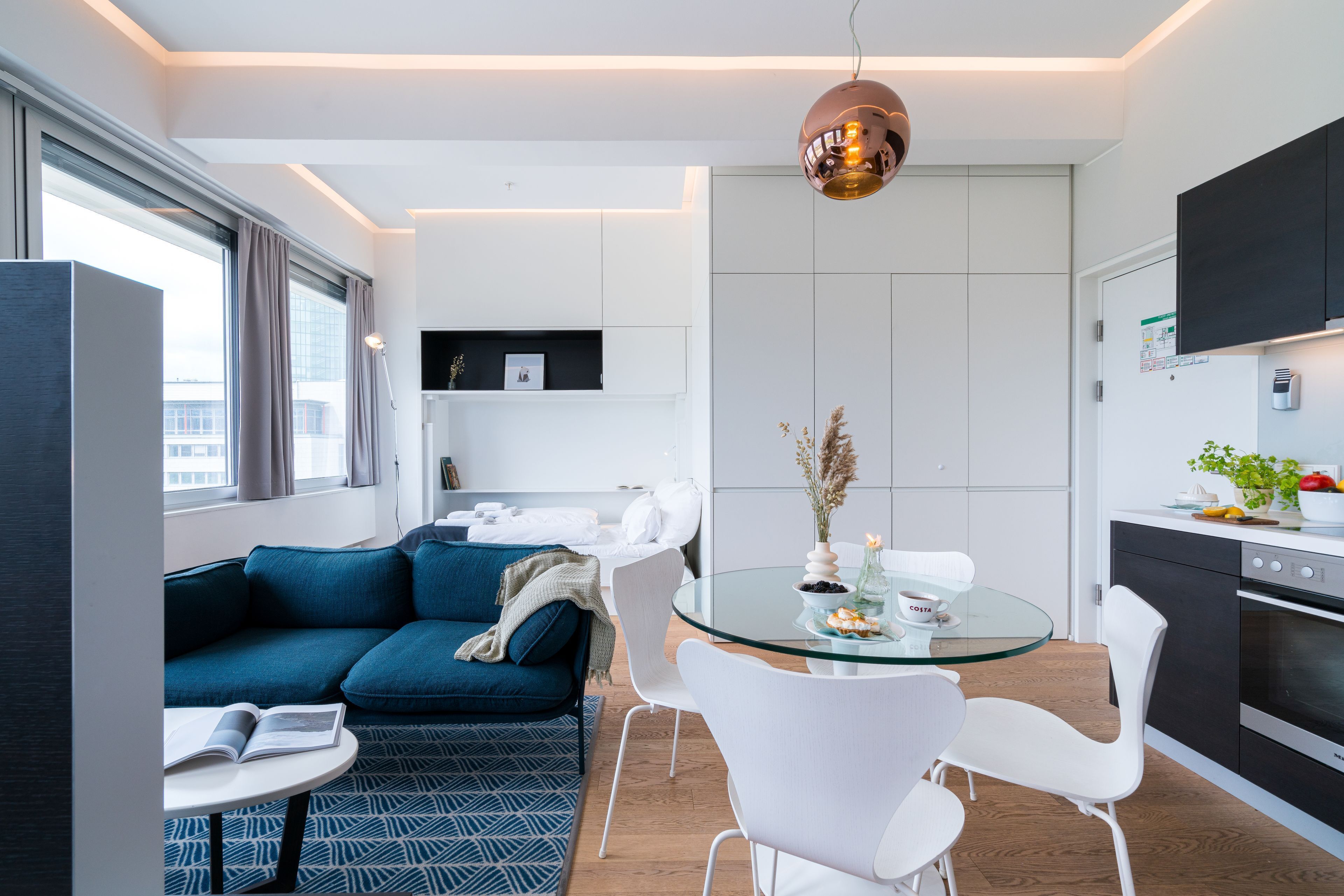 Un salon moderne avec un canapé bleu, des fauteuils blancs, une table basse en verre et un coin cuisine équipé d’une cuisinière et de placards.