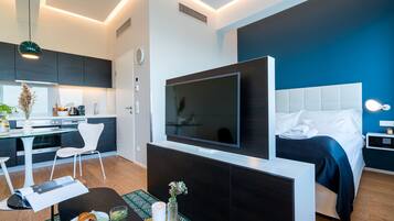 A modern hotel room with a large bed, a flat-screen TV, a dining area with a table and chairs, and a kitchenette.