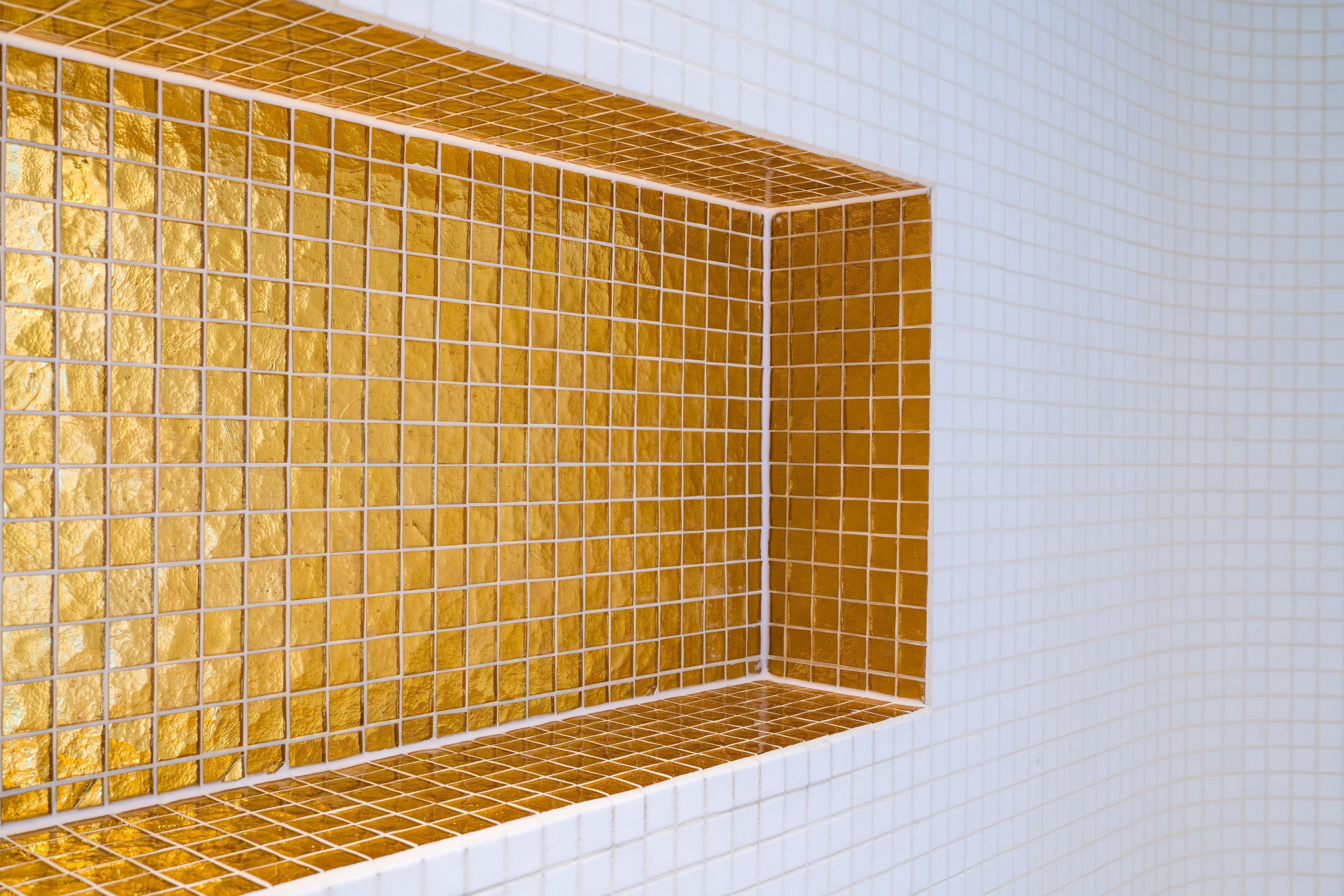 A corner of a room with a white tiled wall and a golden-tiled shelf.