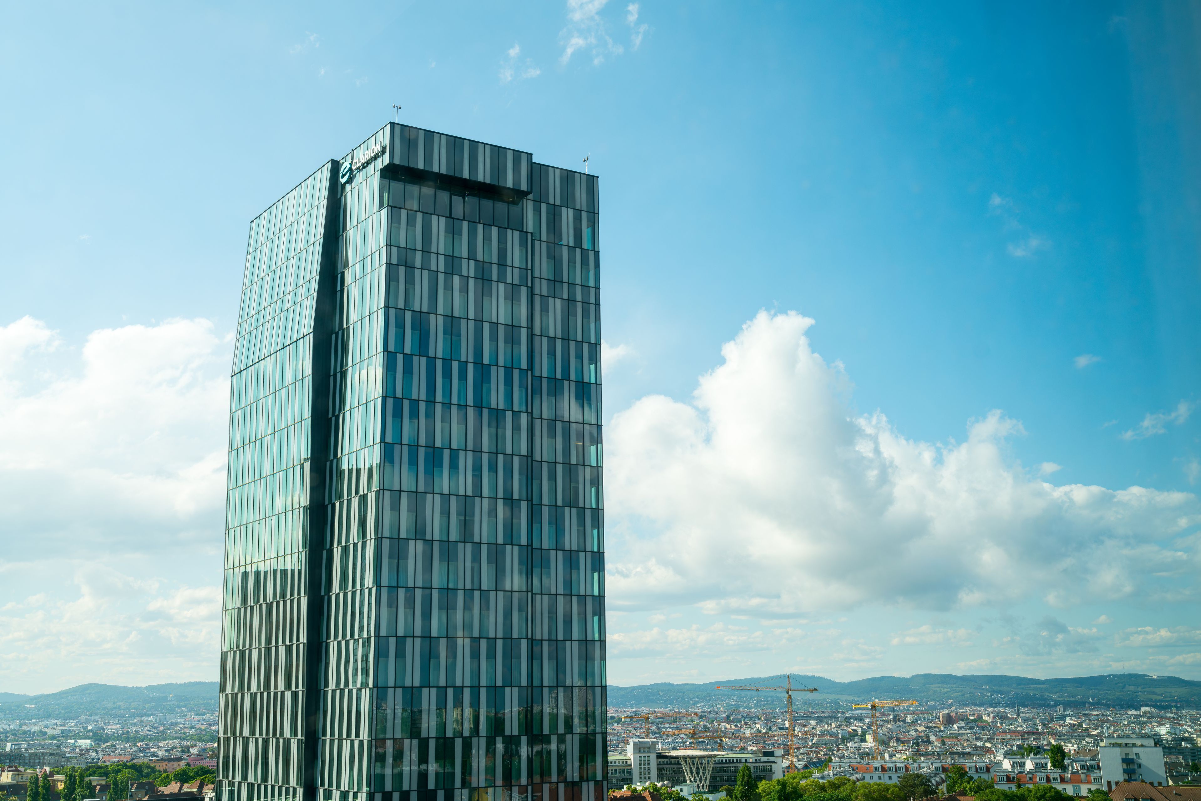 A modern glass skyscraper with a grid-like facade, set against a backdrop of a cityscape and distant mountains.
